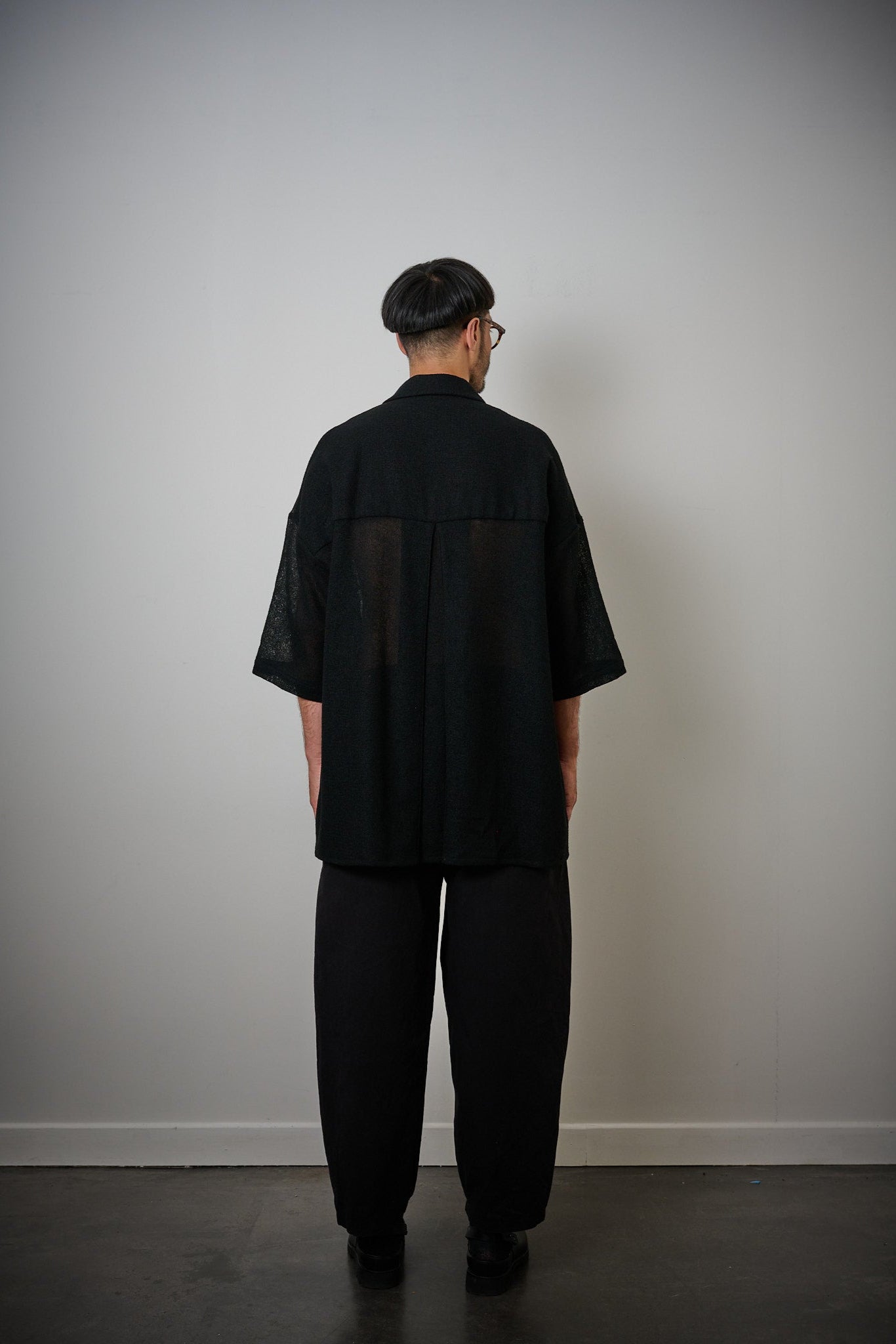 Chemise oversize à col club de la marque Aïdama en crêpe de laine noire légèrement transparente. Portée fermée par un mannequin homme et vu de dos.