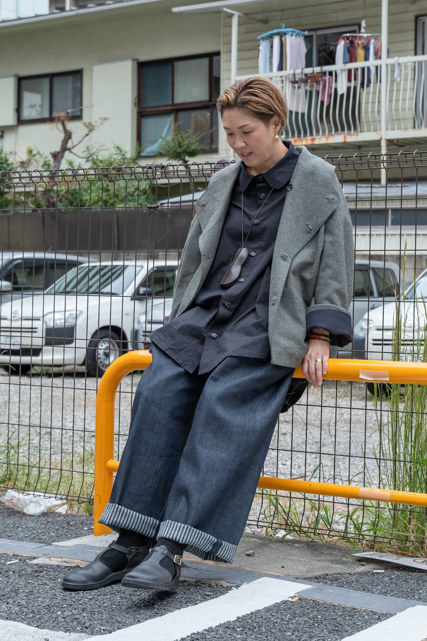 Pantalon droit Aïdama, coupe oversize en denim japonais rayé. Porté par une mannequin femme assis sur une barrière dans une rue japonais et vu de face.