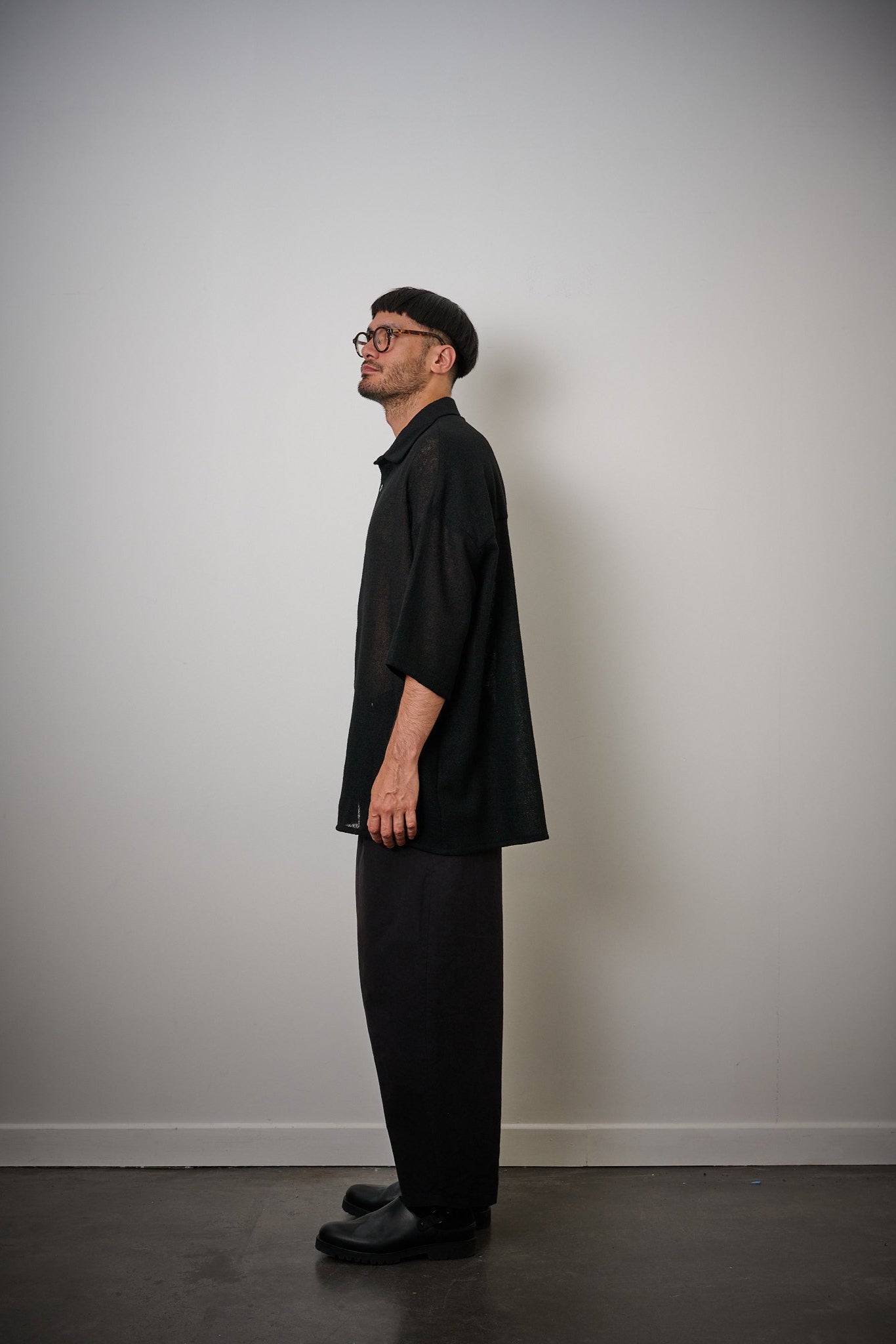 Chemise oversize à col club de la marque Aïdama en crêpe de laine noire légèrement transparente. Portée fermée par un mannequin homme et vue de profil. 