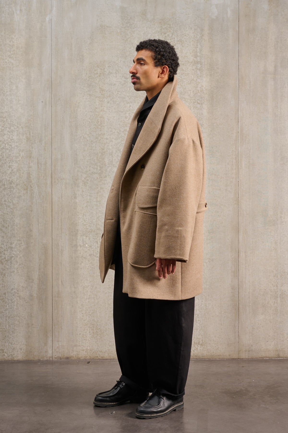 Manteau croisé oversize de la marque Aïdama, modèle Haomi en laine et cachemire beige. Porté ouvert par un mannequin homme et vu de profil.