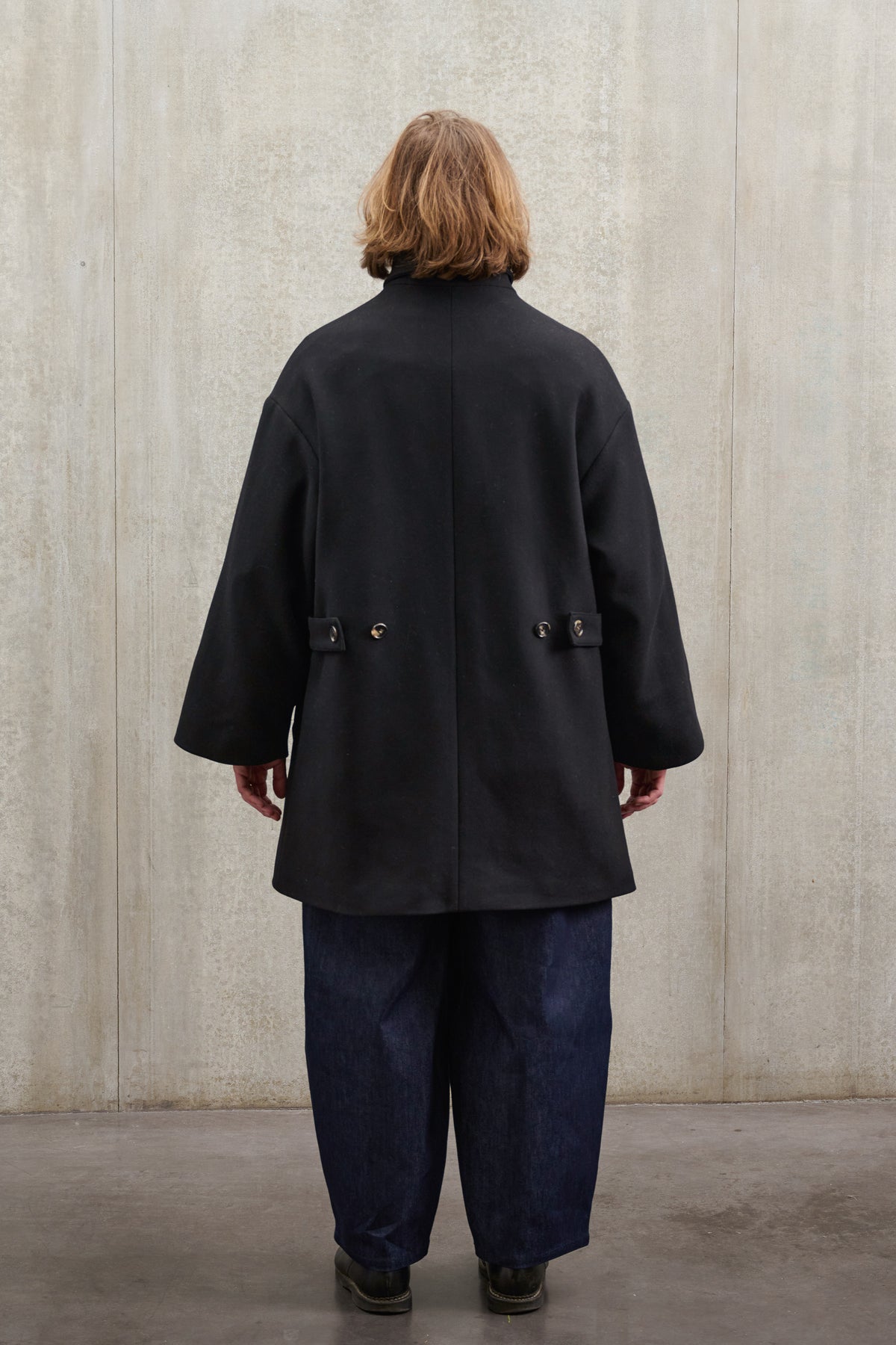 Manteau croisé oversize de la marque Aïdama, modèle Haomi en laine feutrée noire. Porté ouvert par un mannequin homme et vu de dos.