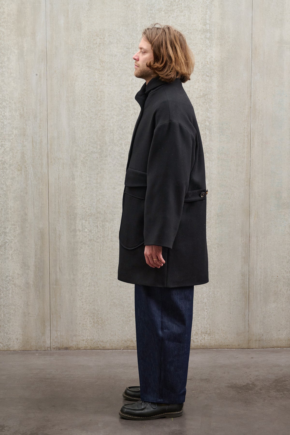 Manteau croisé oversize de la marque Aïdama, modèle Haomi en laine feutrée noire. Porté ouvert par un mannequin homme et vu de profil.