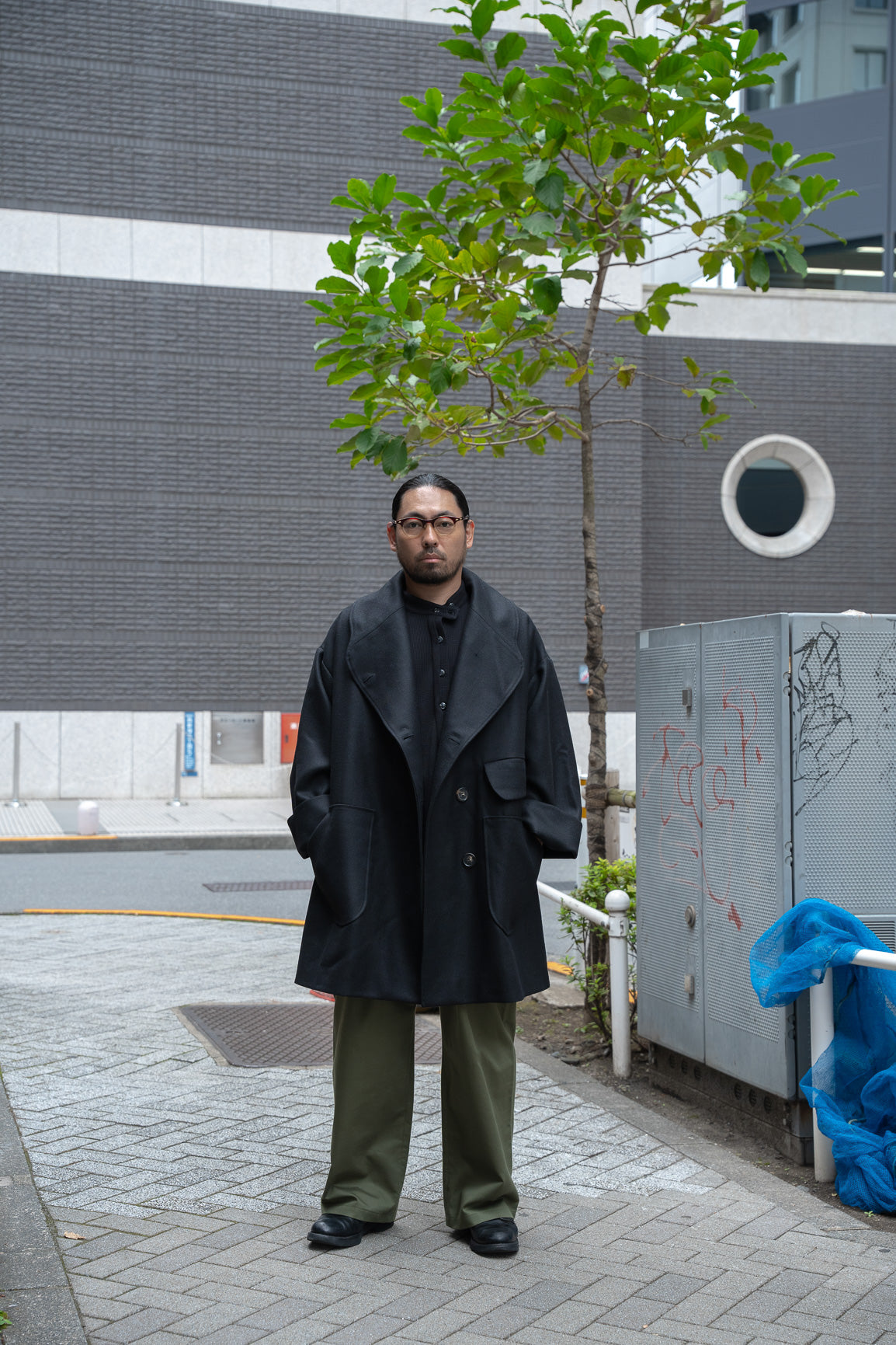 Manteau croisé oversize de la marque Aïdama, modèle Haomi en laine noire. Porté ouvert par un mannequin homme et vu de face en extérieur au Japon.