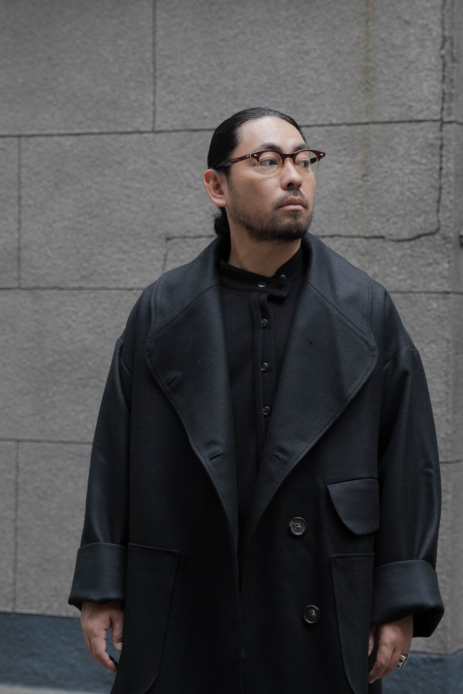 Manteau croisé oversize de la marque Aïdama, modèle Haomi en laine noire. Porté ouvert par un mannequin homme et vu de face en extérieur.