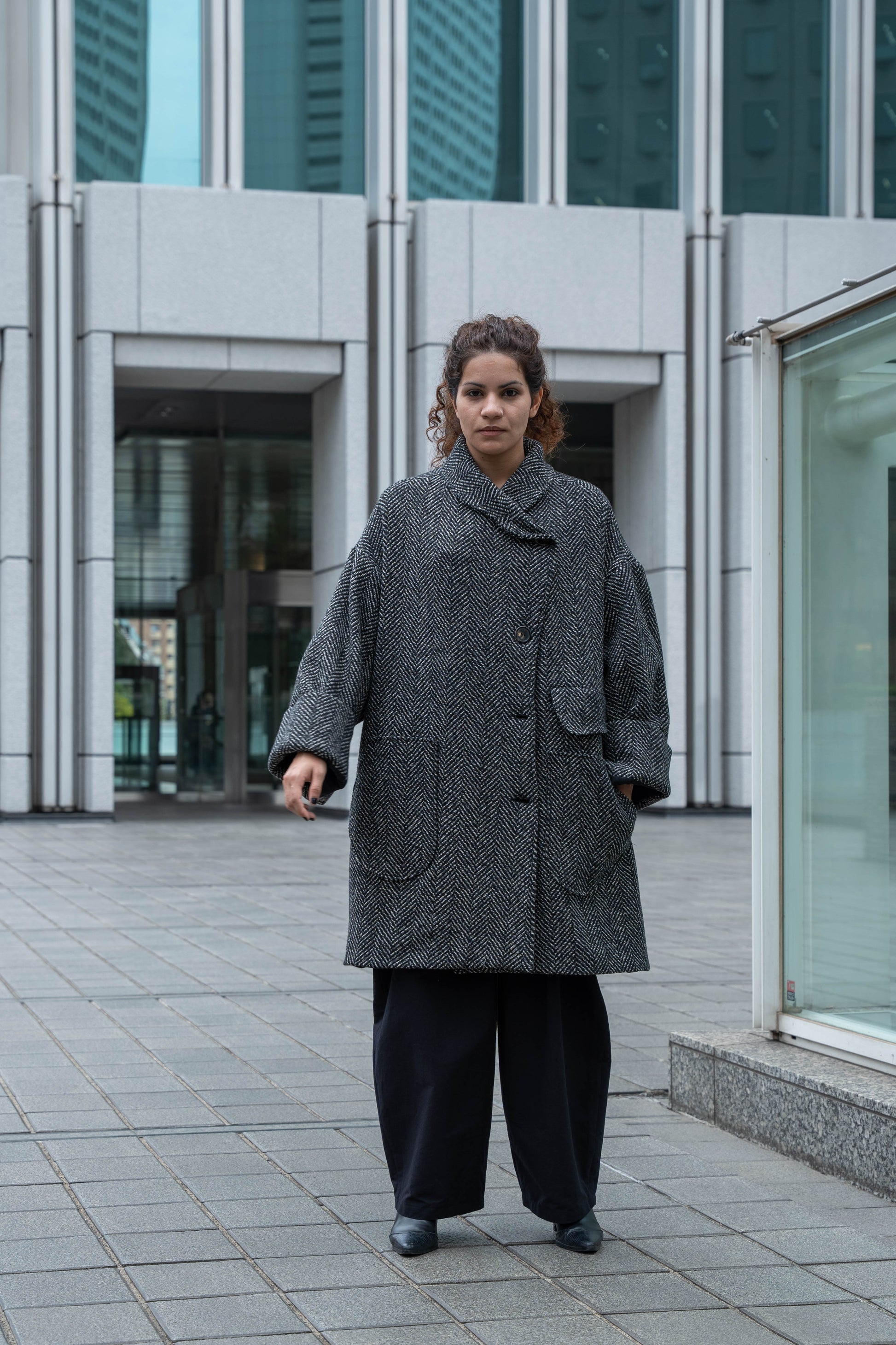 Manteau croisé oversize de la marque Aïdama, modèle Haomi en tweed de laine gris à motif chevrons. Porté fermé par une mannequin femme et vue de face en extérieur.