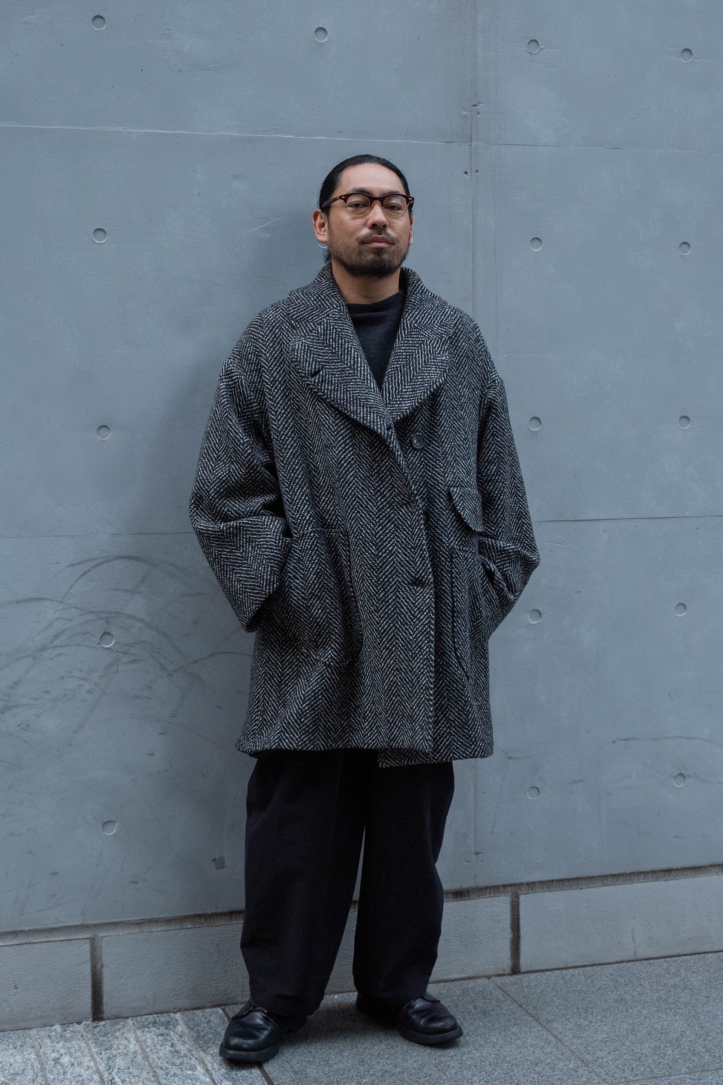 Manteau croisé oversize de la marque Aïdama, modèle Haomi en tweed de laine gris à motif chevrons. Porté fermé par un mannequin homme et vue de face en extérieur.