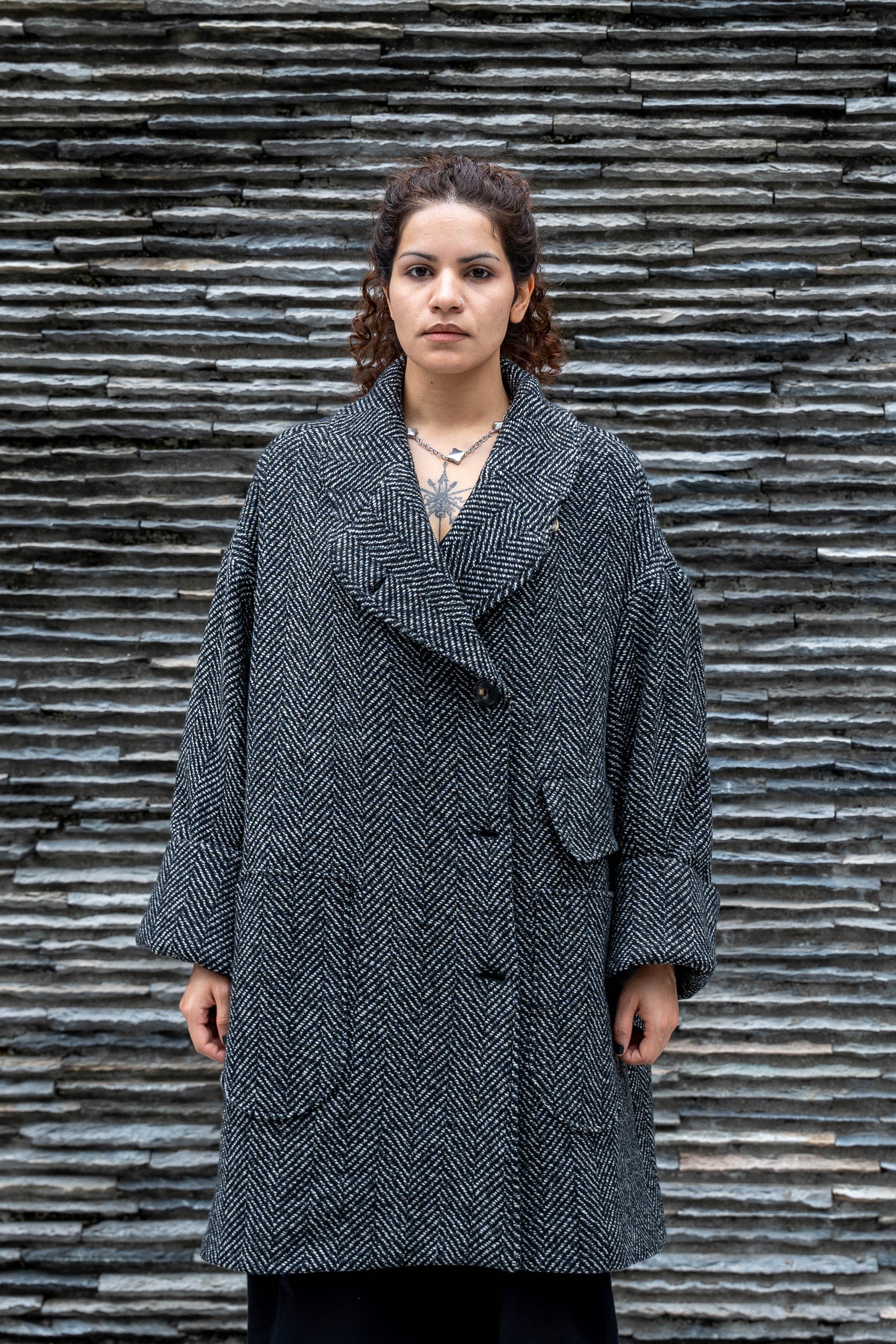 Manteau croisé oversize de la marque Aïdama, modèle Haomi en tweed de laine gris à motif chevrons. Porté fermé par une mannequin femme et vue de face en extérieur.