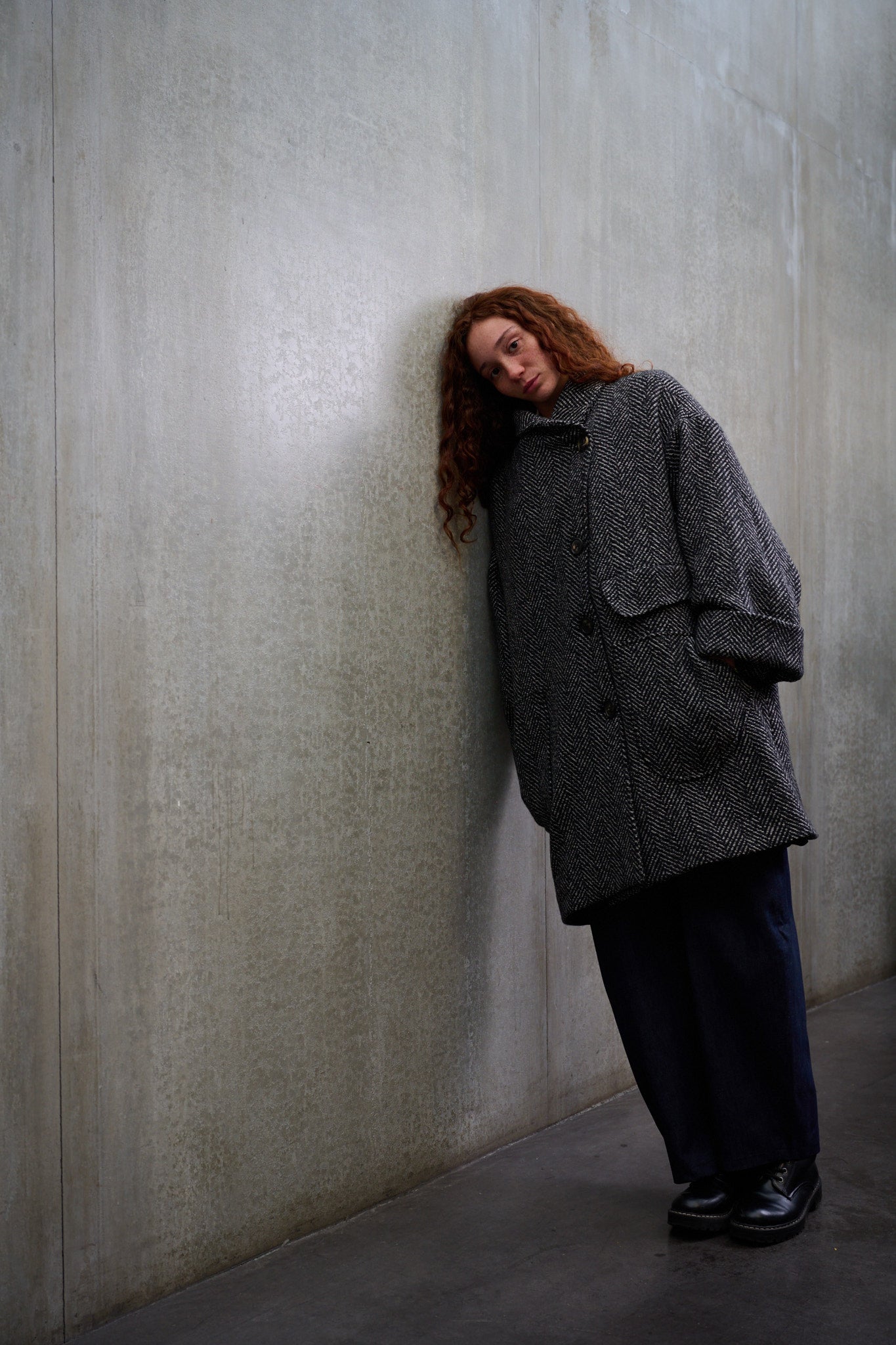 Manteau croisé oversize de la marque Aïdama, modèle Haomi en tweed de laine gris à motif chevrons. Porté fermé par une mannequin femme et vue de face appuyée contre le mur.