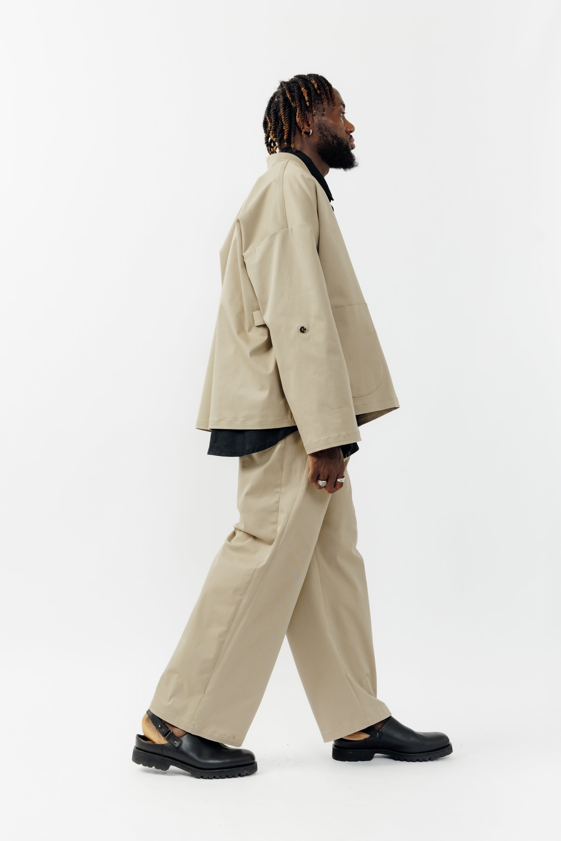 Pantalon droit de la marque Aïdama, coupe ample en gabardine de coton beige avec sangles ajustables à la taille. Porté par un mannequin homme et vu de profil.