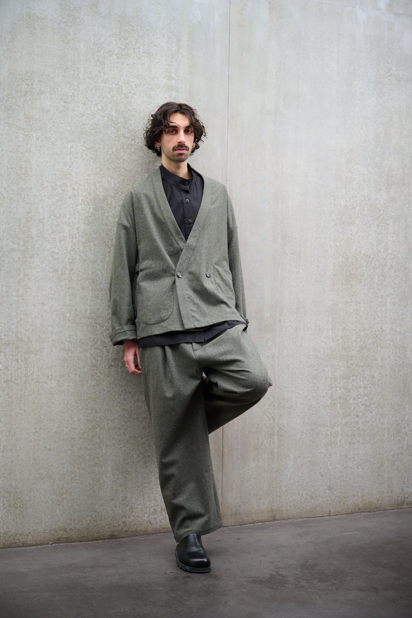 Pantalon droit de la marque Aïdama, coupe large en flanelle de laine grise. Porté par un mannequin homme et vue de face appuyé contre le mur.