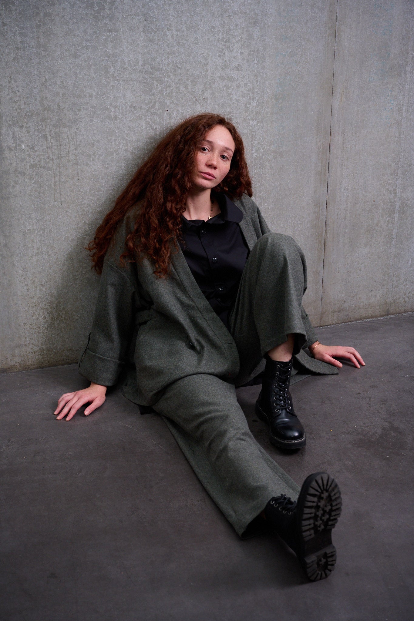 Pantalon droit de la marque Aïdama, coupe large en flanelle de laine grise. Porté par une mannequin femme et vue de face assise par terre, contre le mur.