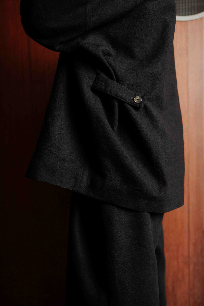 Veste oversize à col arrondi de la marque Aïdama, modèle Gunbai en laine et lin noir Portée fermée par un mannequin homme et vue de profil zoomée sur la sangle de réglage.