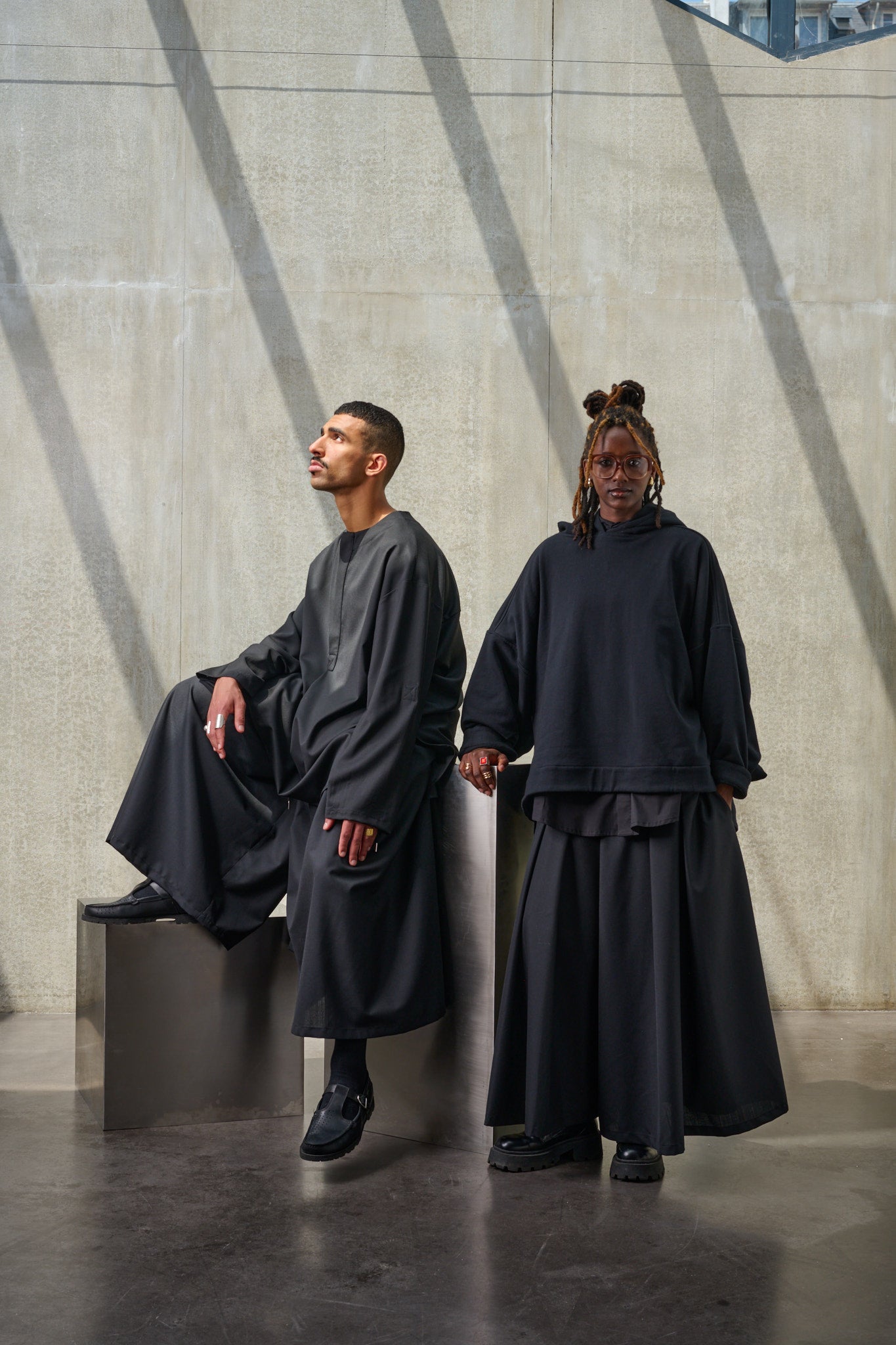 Jupe culotte oversize de la marque Aïdama, modèle Hakama en laine froide noire, portée par un modèle homme assis et un modèle femme debout.