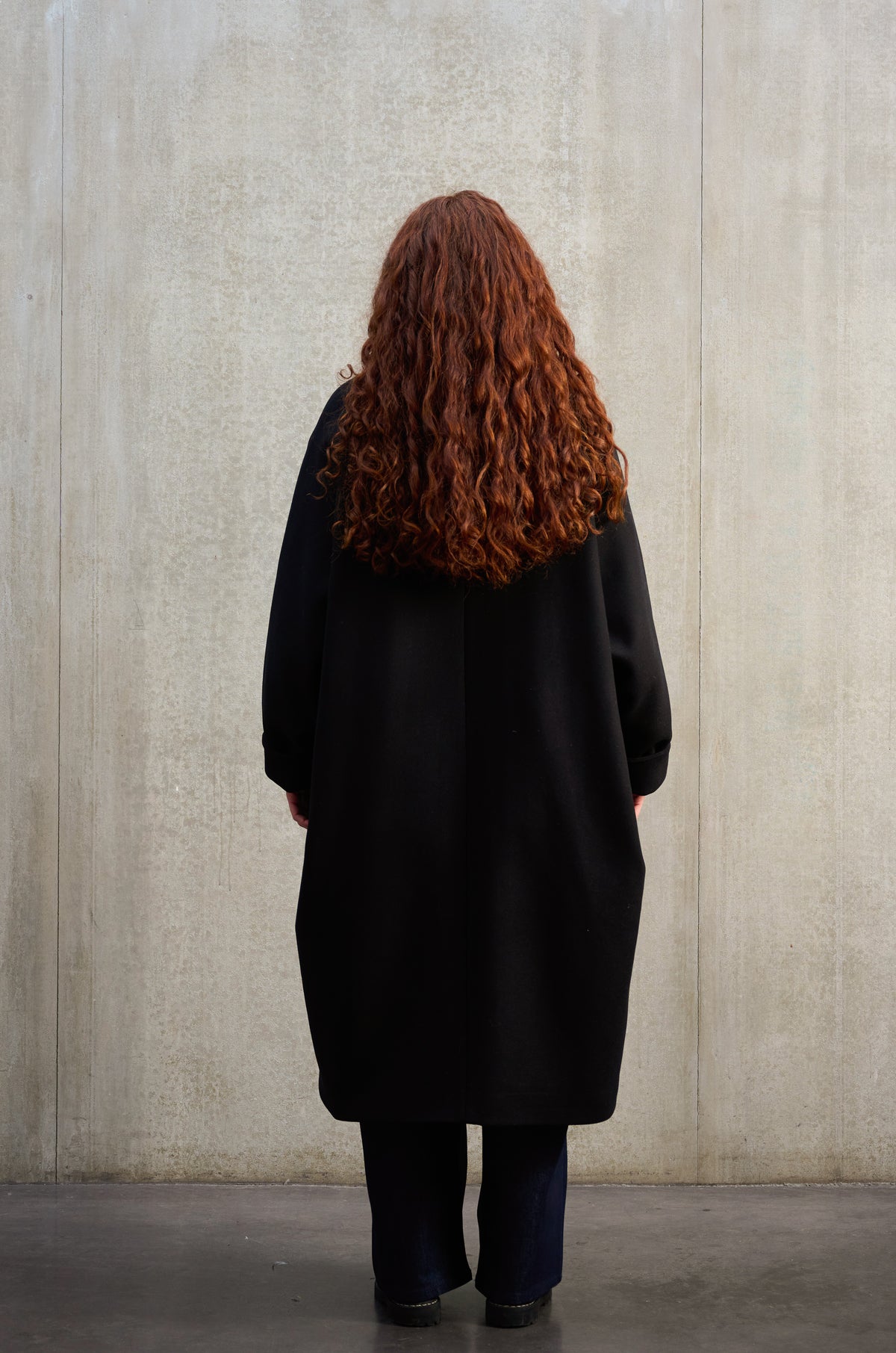 Manteau oversize à manches dolmans de la marque Aïdama, modèle Tanzen en laine feutrée noir. Porté ouvert par une mannequin femme et vu de dos.
