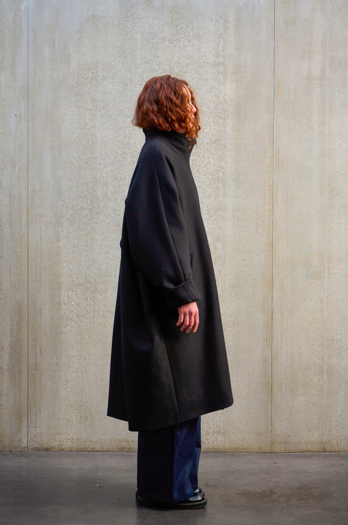 Manteau oversize à manches dolmans de la marque Aïdama, modèle Tanzen en laine feutrée noir. Porté ouvert par une mannequin femme et vu de profil.