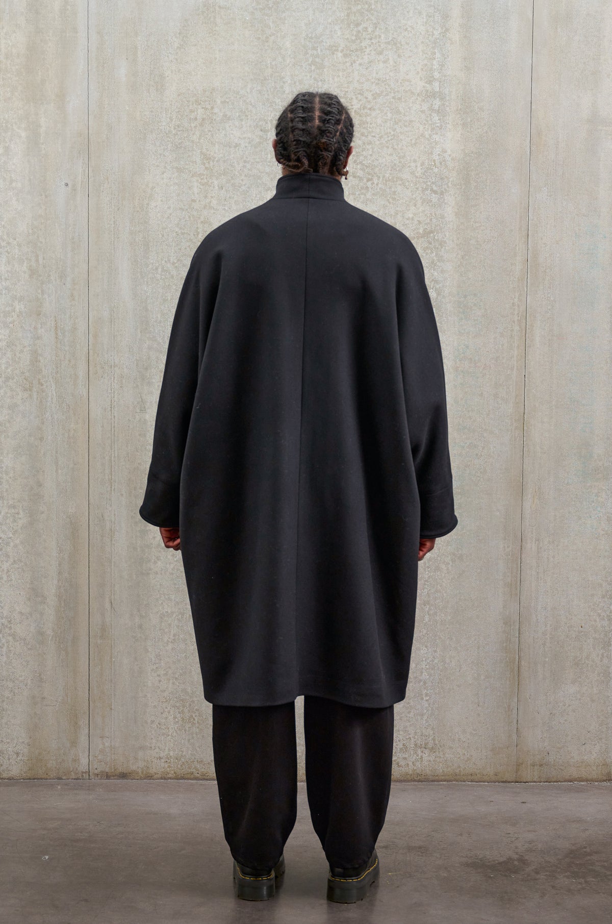 Manteau oversize à manches dolmans de la marque Aïdama, modèle Tanzen en laine feutrée noir. Porté fermé par un mannequin homme et vu de dos.