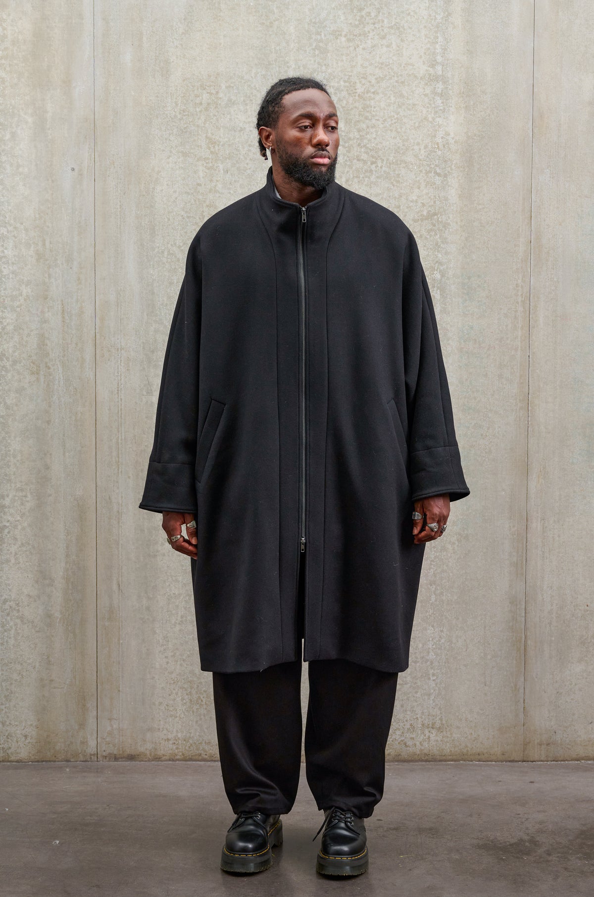 Manteau oversize à manches dolmans de la marque Aïdama, modèle Tanzen en laine feutrée noir. Porté fermé par un mannequin homme et vu de face.