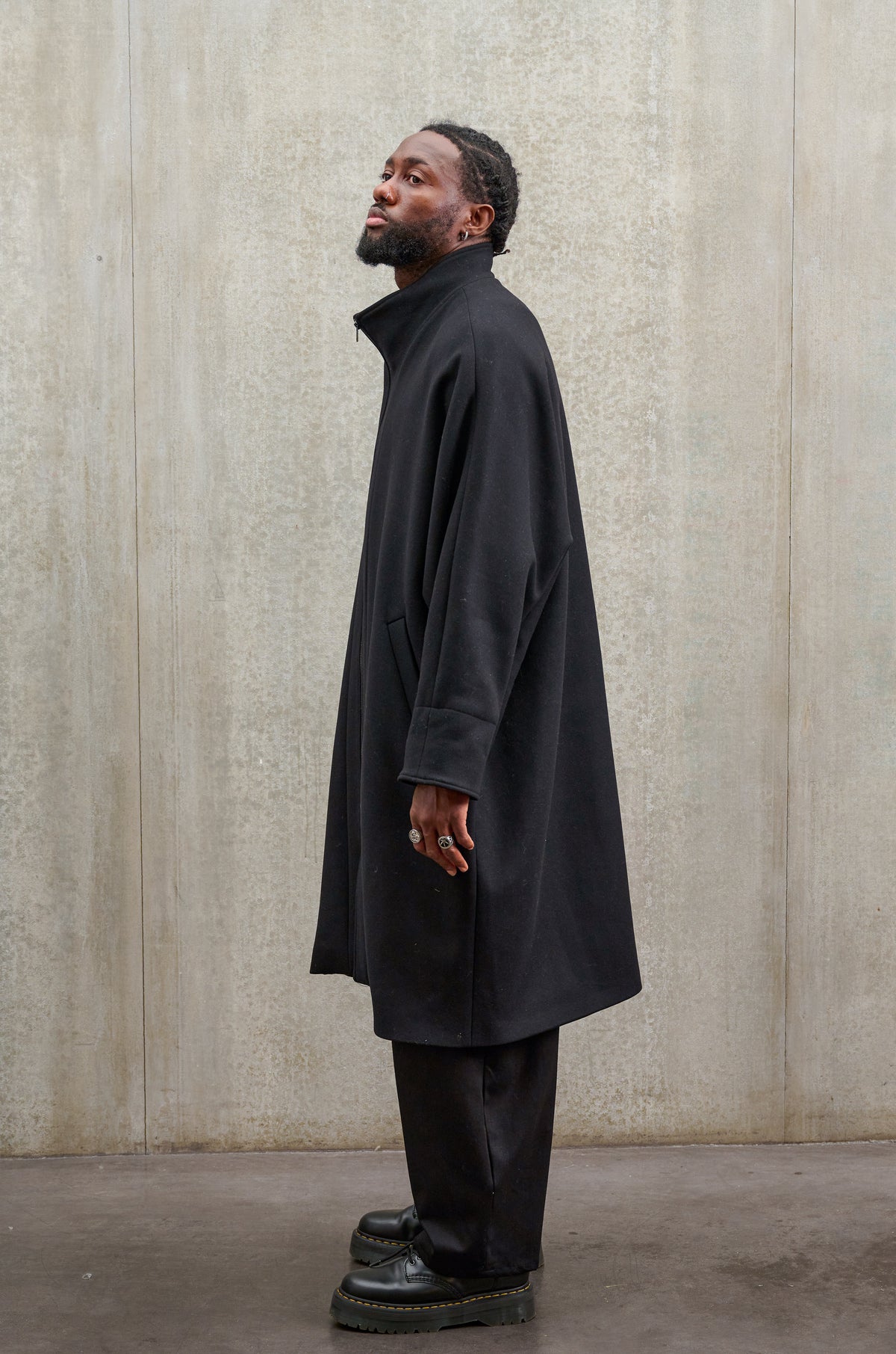 Manteau oversize à manches dolmans de la marque Aïdama, modèle Tanzen en laine feutrée noir. Porté fermé par un mannequin homme et vu de profil