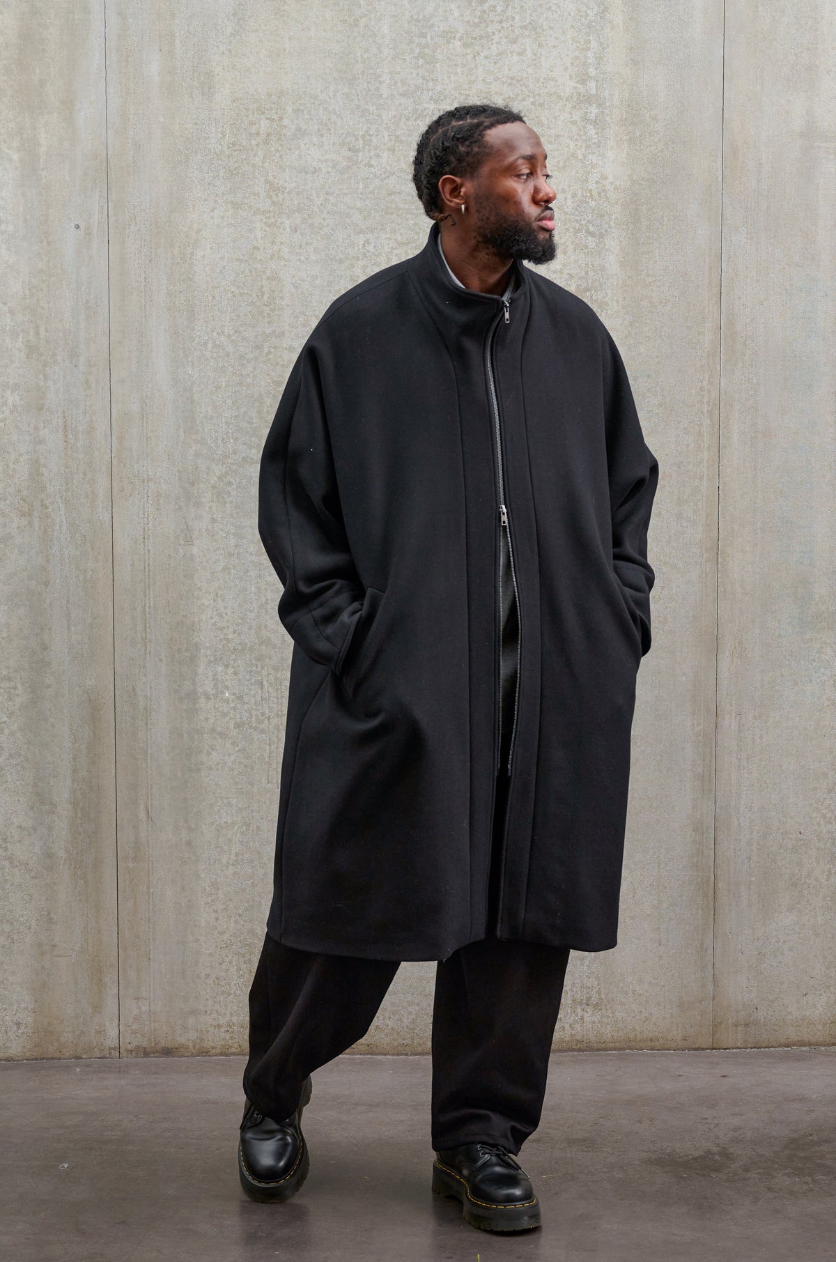Manteau oversize à manches dolmans de la marque Aïdama, modèle Tanzen en laine feutrée noir. Porté fermé par un mannequin homme et vu de face.