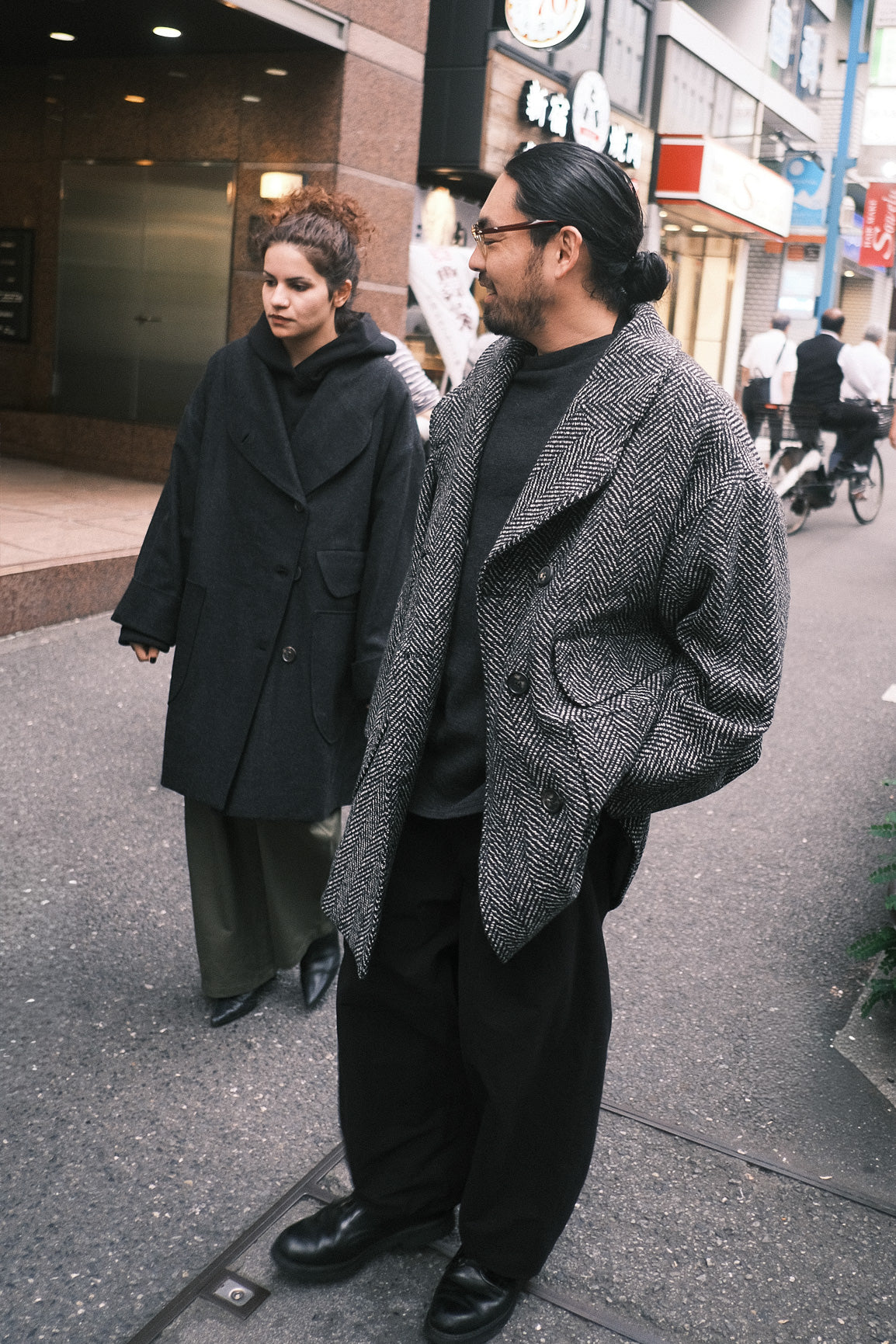 Manteau croisé oversize de la marque Aïdama, modèle Haomi en tweed de laine gris à motif chevrons. Porté ouvert par un mannequin homme et vue de face en extérieur dans une rue au Japon.