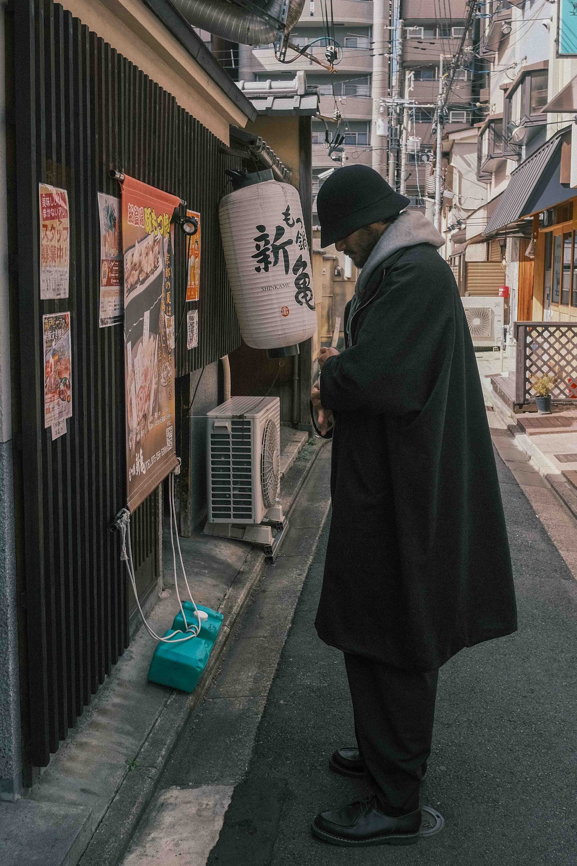 Manteau long à manches dolmans de la marque Aïdama, modèle Tanzen en laine noire. Porté par un mannequin homme et vu de profil en extérieur au Japon. 
