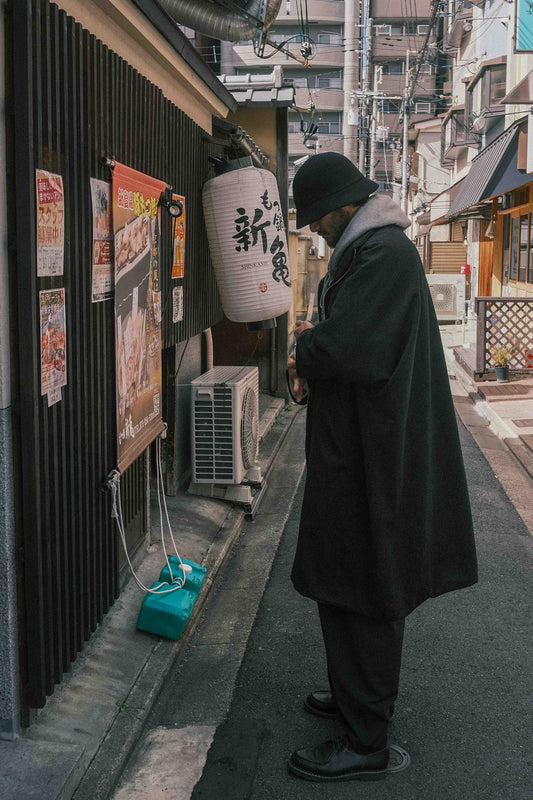 Manteau long à manches dolmans de la marque Aïdama, modèle Tanzen en laine noire. Porté par un mannequin homme et vu de profil en extérieur au Japon. 