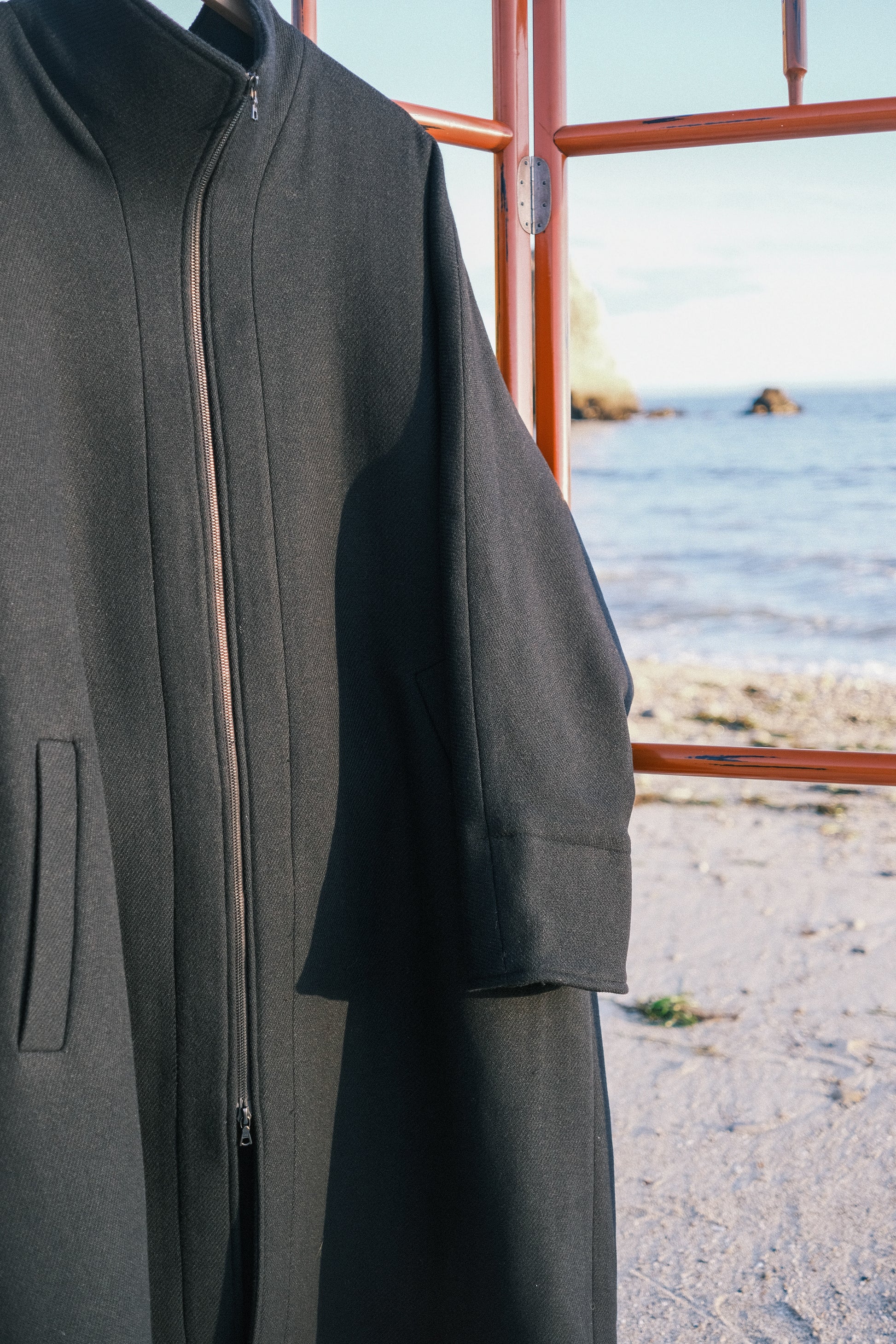 Manteau long à manches dolmans de la marque Aïdama, modèle Tanzen en laine noire. Vu sur cintre en extérieur sur une plage.