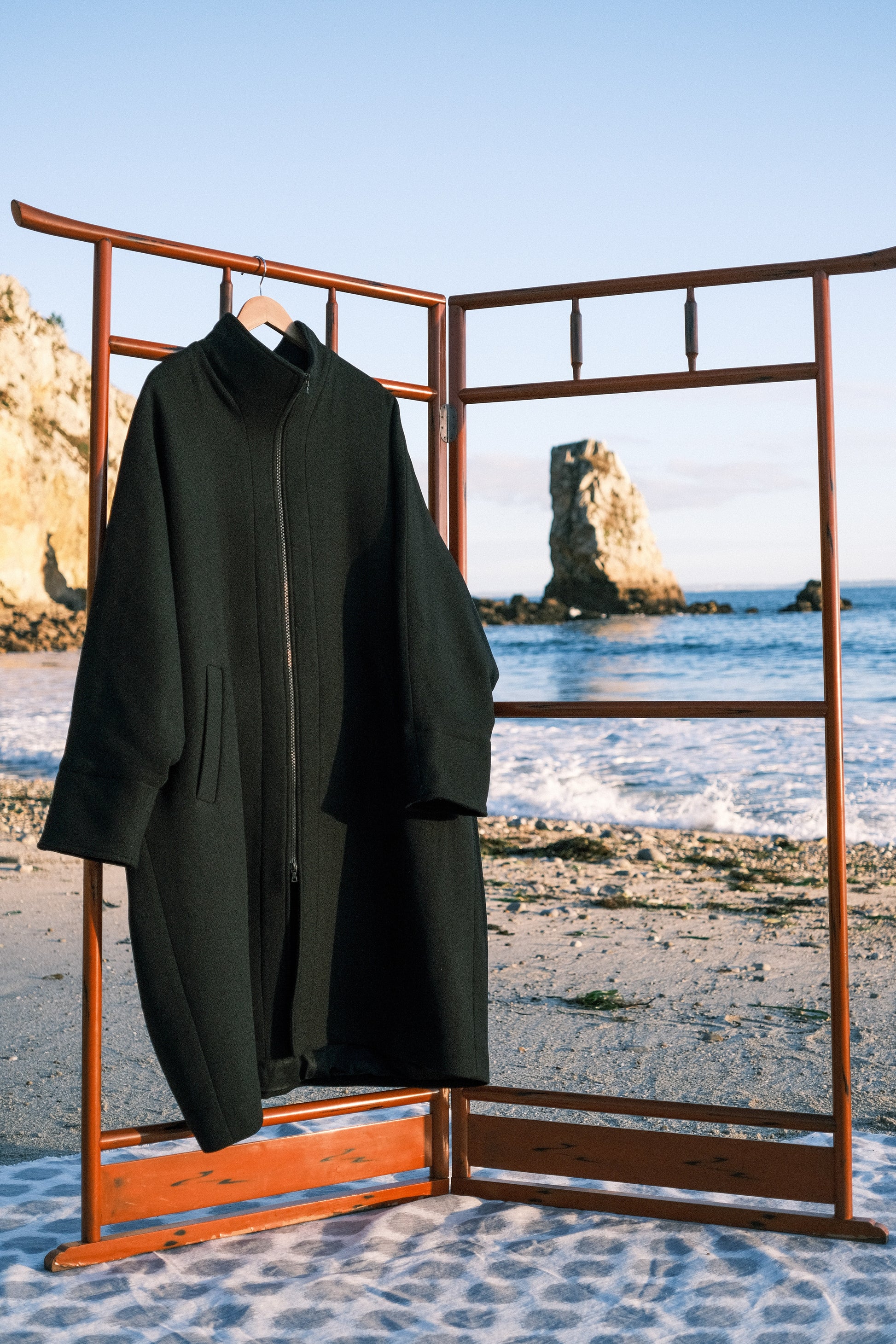 Manteau long à manches dolmans de la marque Aïdama, modèle Tanzen en laine noire. vu de face sur cintre en extérieur sur une plage.