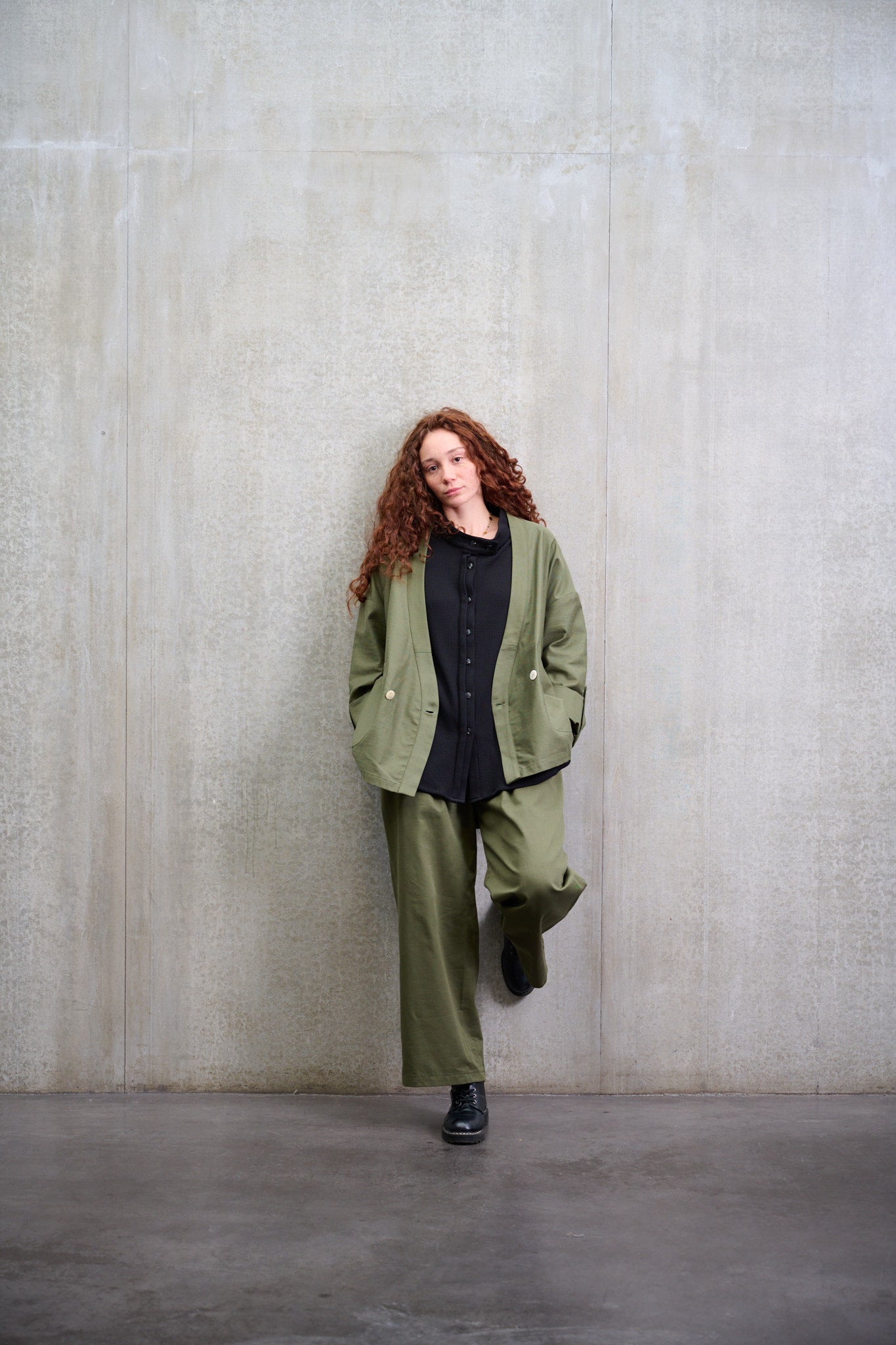 Pantalon droit à coupe ample en coton vert avec sangles de réglages de la marque Aïdama. Porté par une mannequin et vu de face appuyé contre le mur.