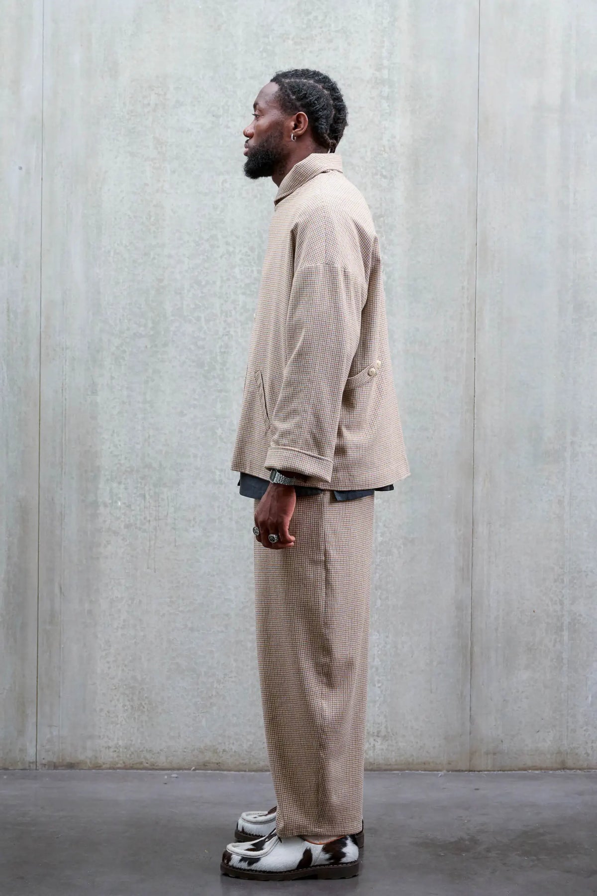 Pantalon circulaire unisexe de la marque Aïdama, coupe ballon en laine marron à motif pied de poule. Porté par un mannequin homme et vu de profil.