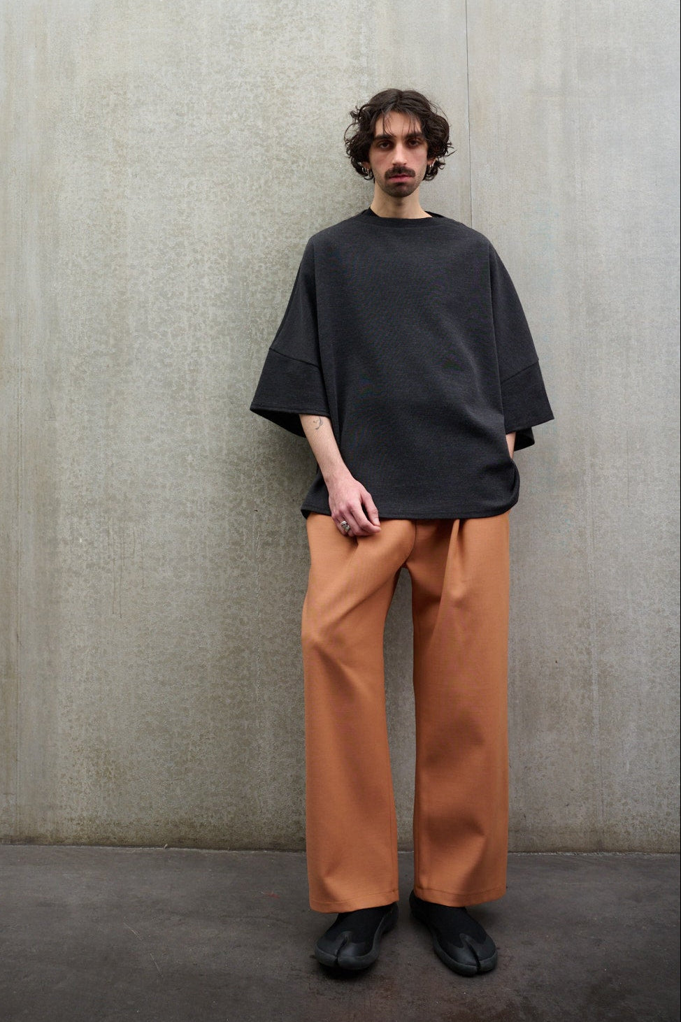 Pantalon droit de la marque Aïdama coupe oversize en laine et coton orange. Porté par un mannequin homme, vu de face appuyé contre le mur.