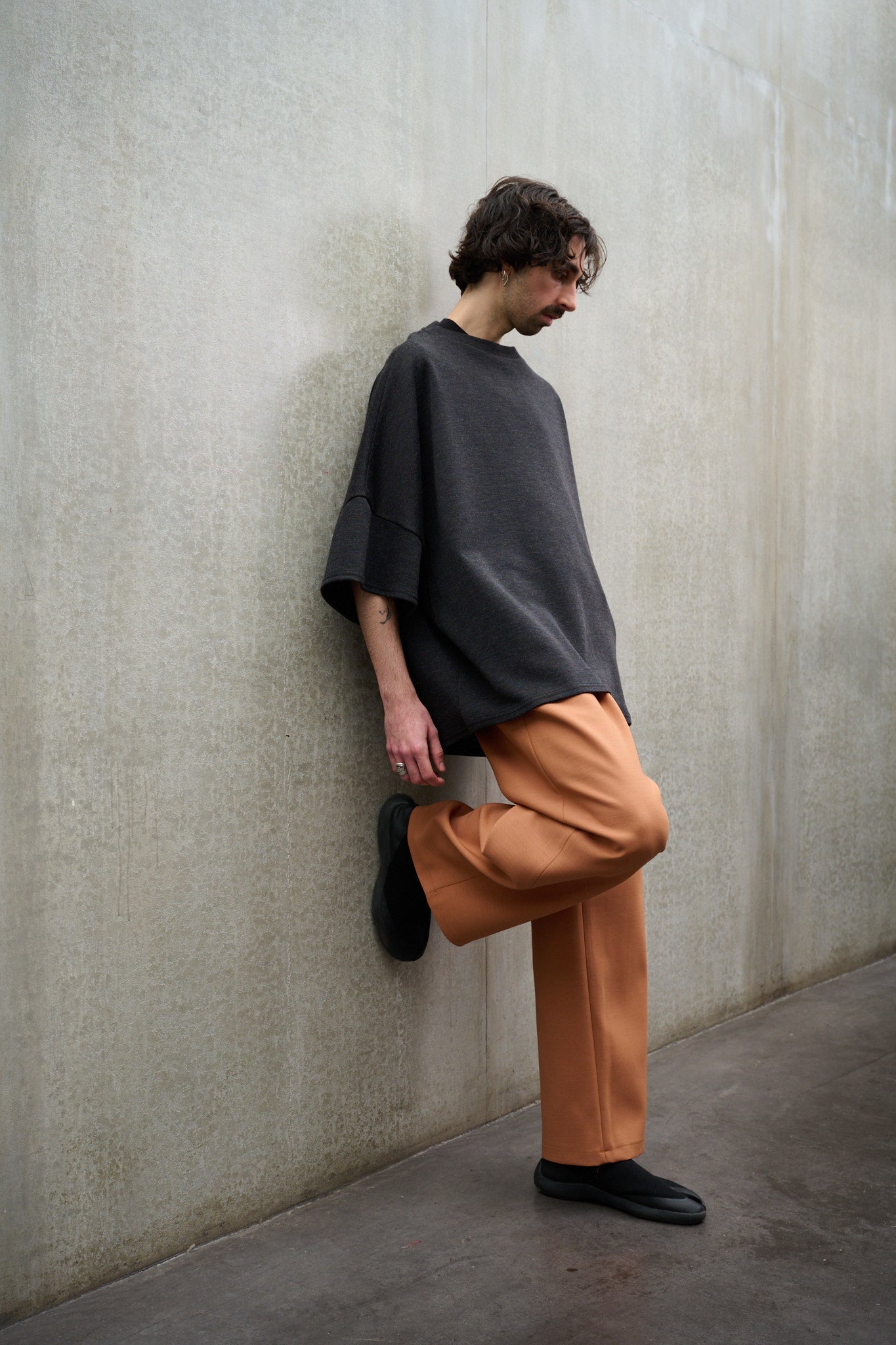 Pantalon droit de la marque Aïdama coupe oversize en laine et coton orange. Porté par un mannequin homme, vu de profil appuyé contre le mur.