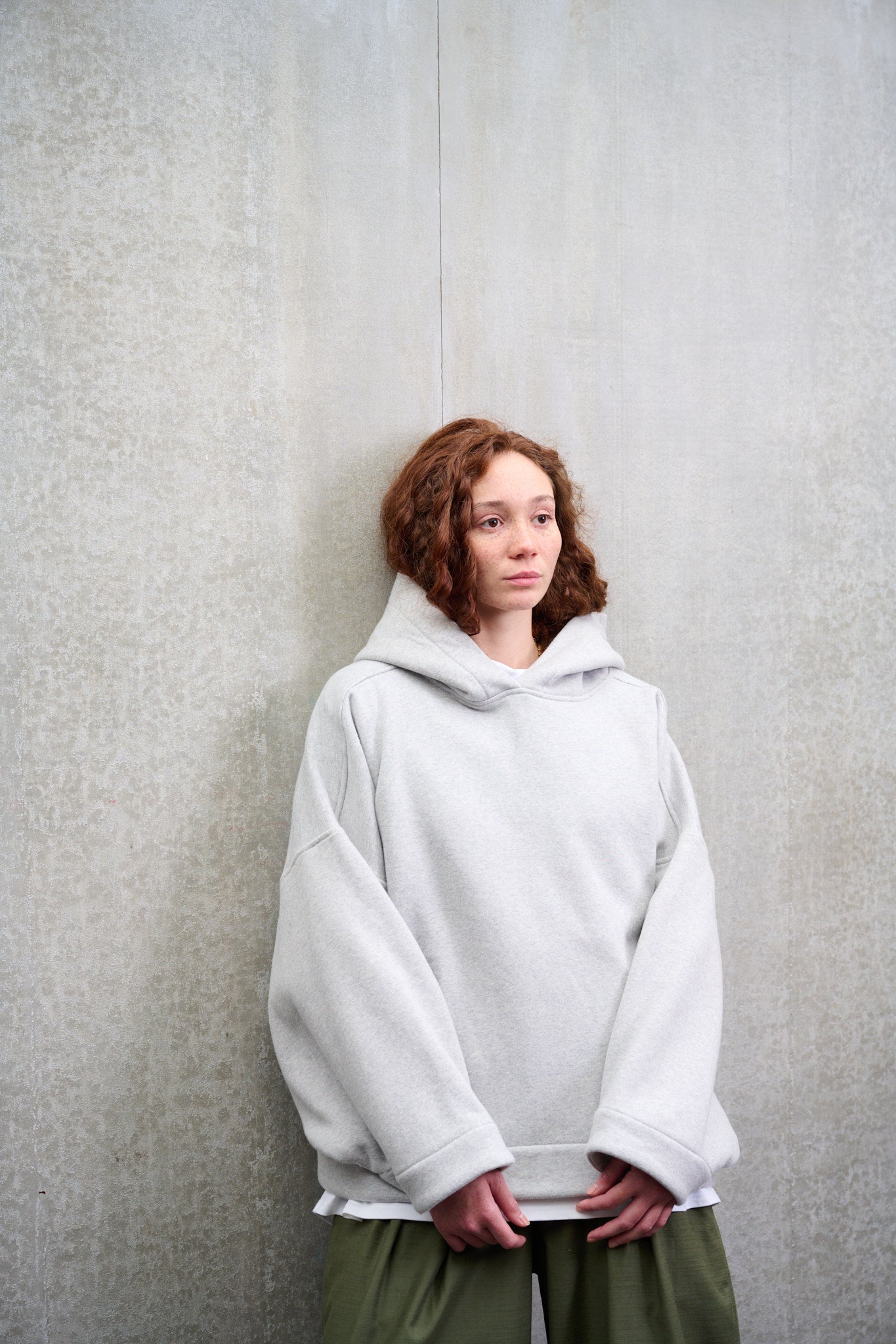 Sweat à capuche oversize en molleton de coton gris chiné Mino de la marque Aïdama. Porté par une mannequin femme et vu de face adossée contre le mur.