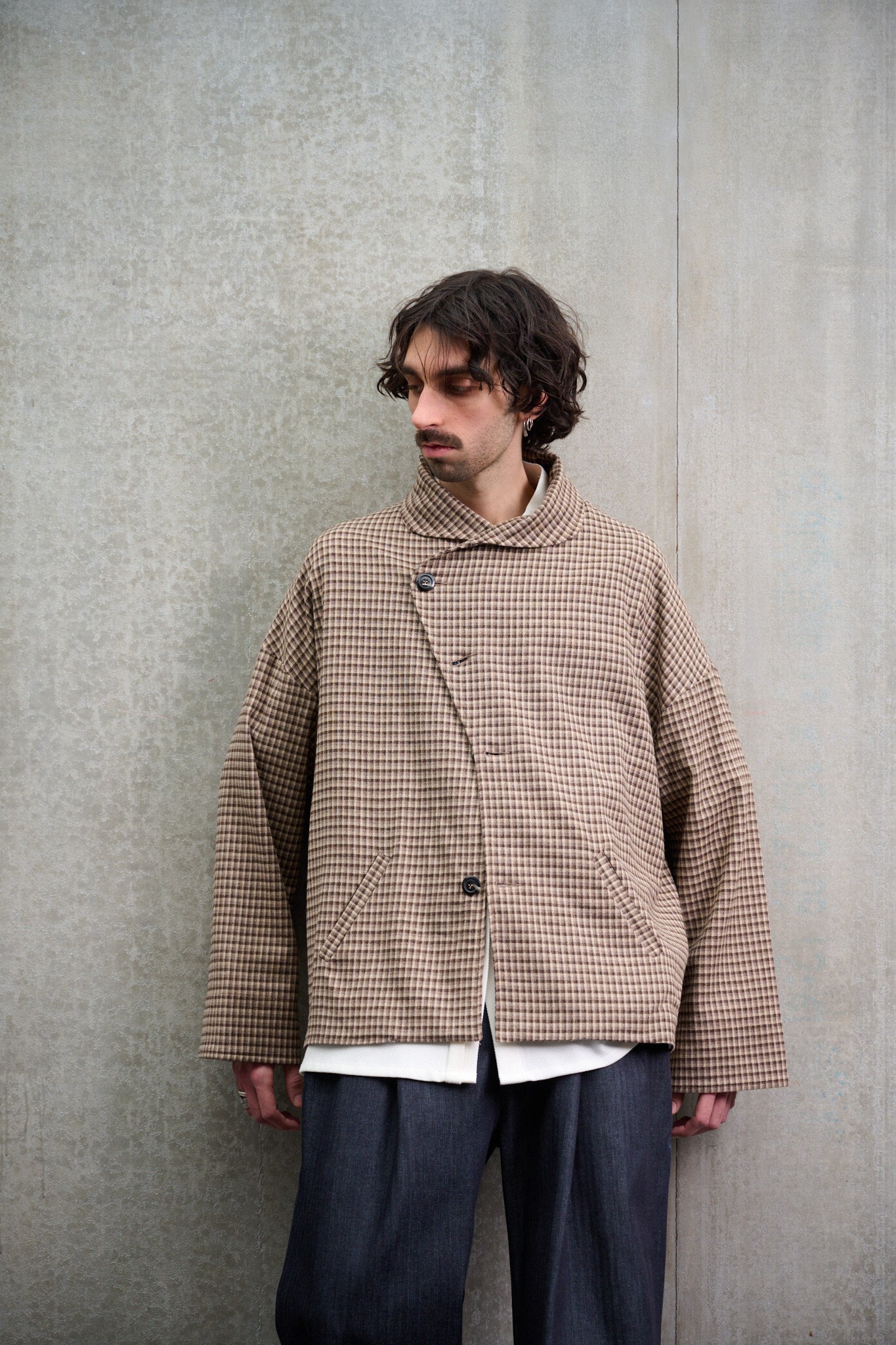 Veste oversize à col arrondi de la marque Aïdama, modèle Gunbai en laine marron à motif Vichy. Portée fermée par un mannequin homme et vue de face appuyé contre un mur.