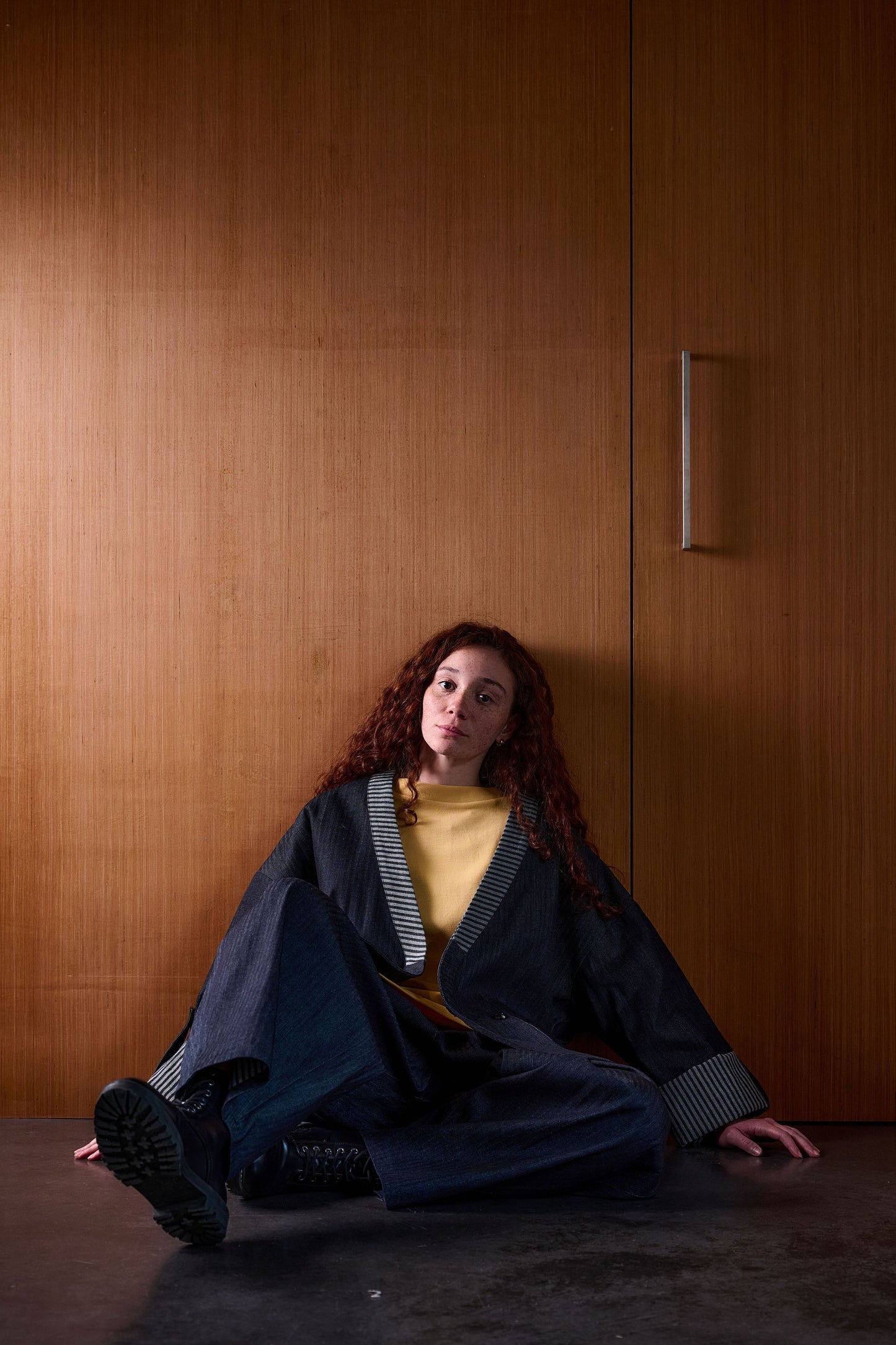 Veste croisée à col kimono Aïdama modèle samue en denim japonais brut. Portée ouverte par une mannequin femme assise et adossée à une porte.